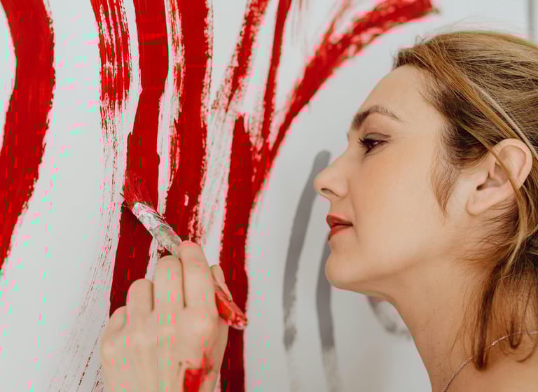 Close-up de uma mulher aplicando pinceladas de tinta vermelha vibrante em uma superfície branca.