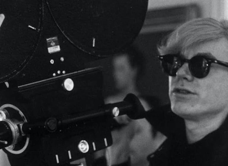 Andy Warhol segurando uma câmera de filme antiga, usando óculos escuros preta
