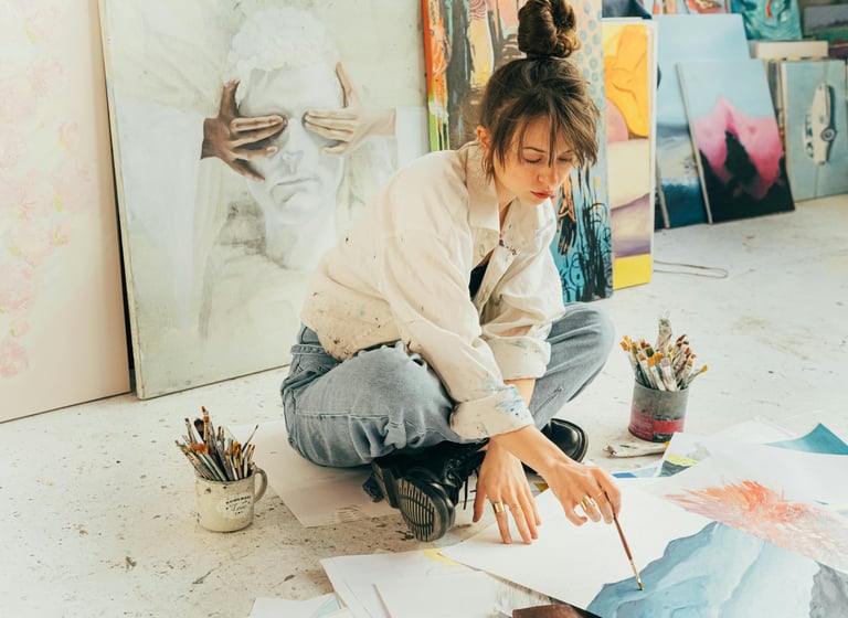 Jovem artista sentada no chão rodeada por pinturas, trabalhando em uma aquarela.