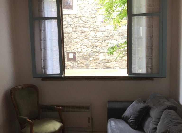 Salon rustique avec canapé gris, fauteuil vert et fenêtre ouverte donnant sur l’église de Quillan