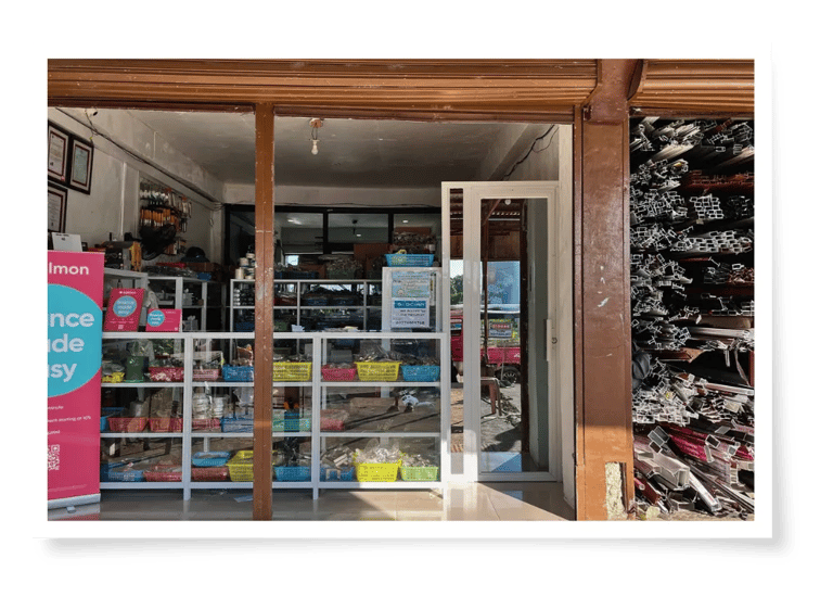 A view of a hardware store entrance with shelves of colorful supplies and metal materials stacked outside. Sunlight brightens