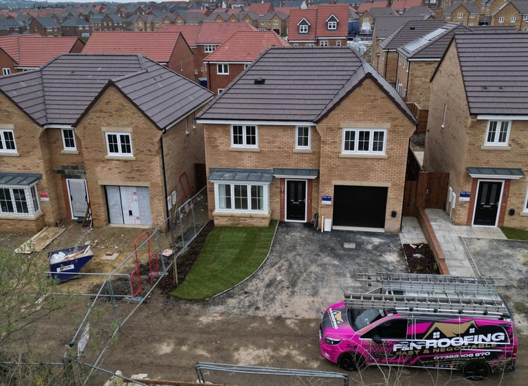A roofing company van parked in front of a new build brick house on a modern residential construction site.