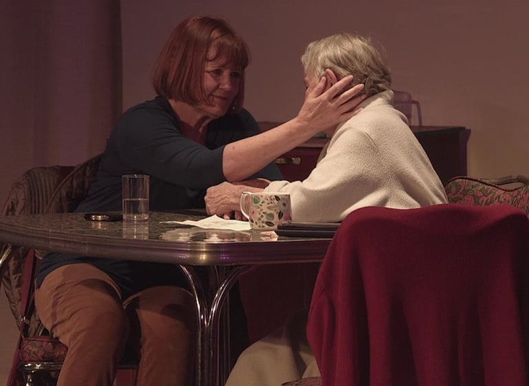 An adult daughter caressing her mature mother's cheek as they sit chatting at a dining table.