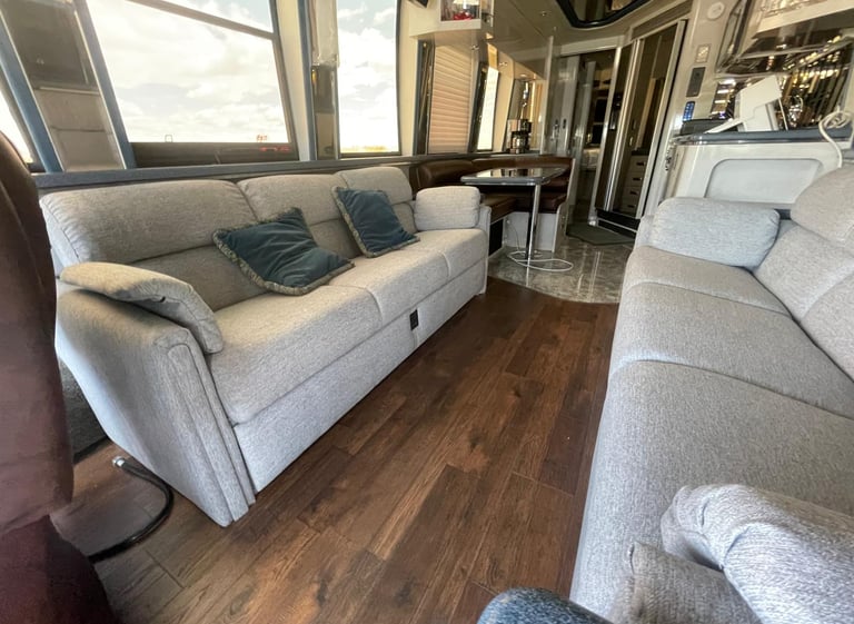 Luxury RV interior with grey fabric sofas, dark wood flooring, and a dinette area in a modern motorhome.