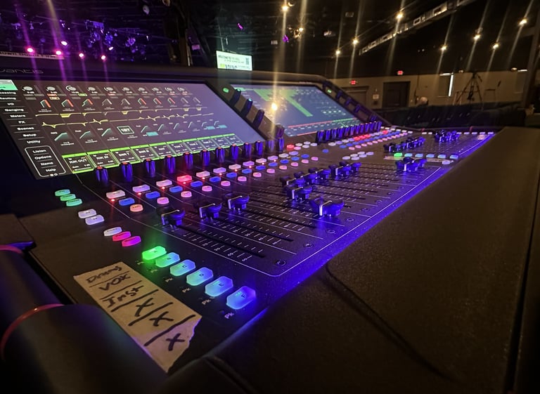 Professional digital audio mixing console with glowing LED buttons and faders in a dark concert venue.