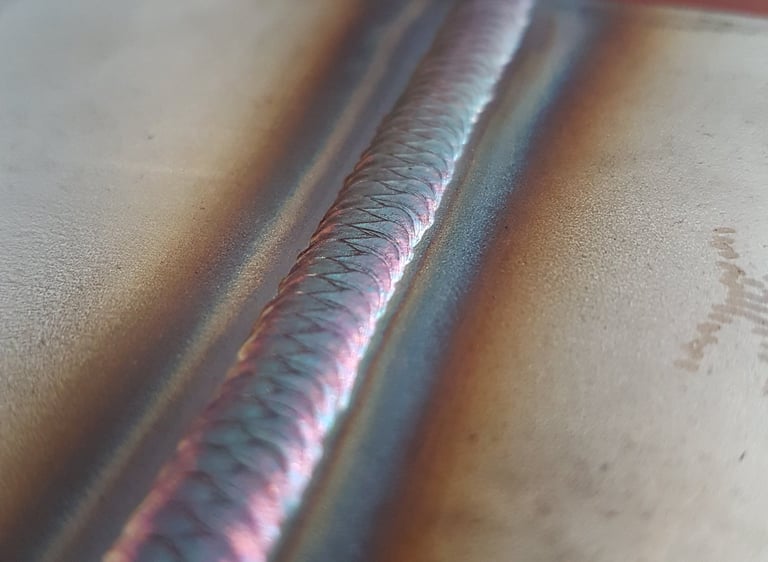 A precise TIG weld bead on stainless steel showing colorful heat tint and uniform ripples.