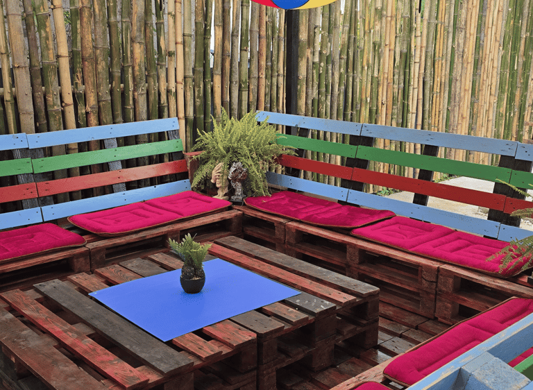 Outdoor patio lounge with colorful umbrella ceiling, wooden pallet furniture, and bamboo privacy fence.