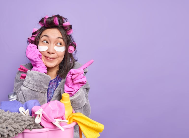 A woman in hair rollers and eye patches wearing rubber gloves with cleaning supplies against a purple background.