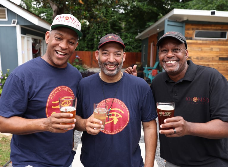 Three smiling men toast with glasses of craft beer while wearing Khonso Brewing apparel outdoors.