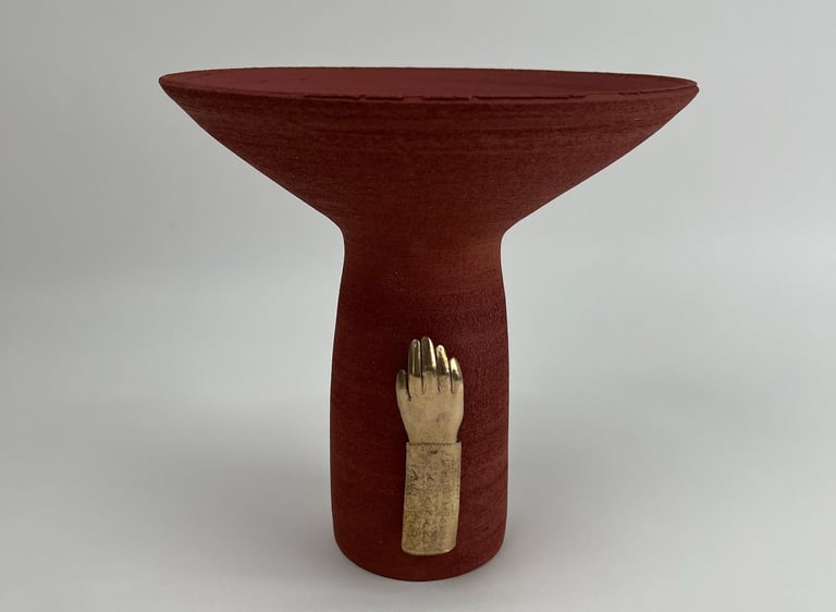 A tall thrown red ceramic vessel, wide at the top. A cast bronze hand is fixed on its base.