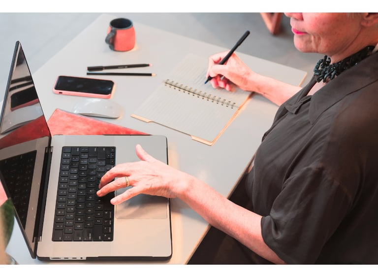 Uma mulher consulta o notebook aberto em cima da mesa enquanto faz anotações no caderno ao lado.