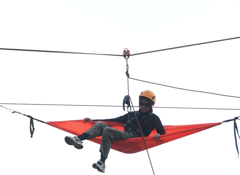 A person wearing a safety helmet relaxes in an orange hammock suspended high on a zip line cable.