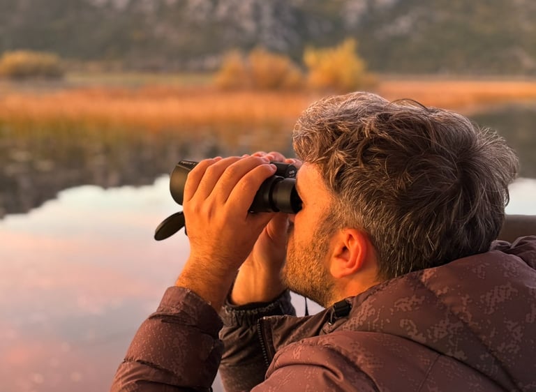 A man uses binoculars for birdwatching over a lake with scenic mountain views at sunset.