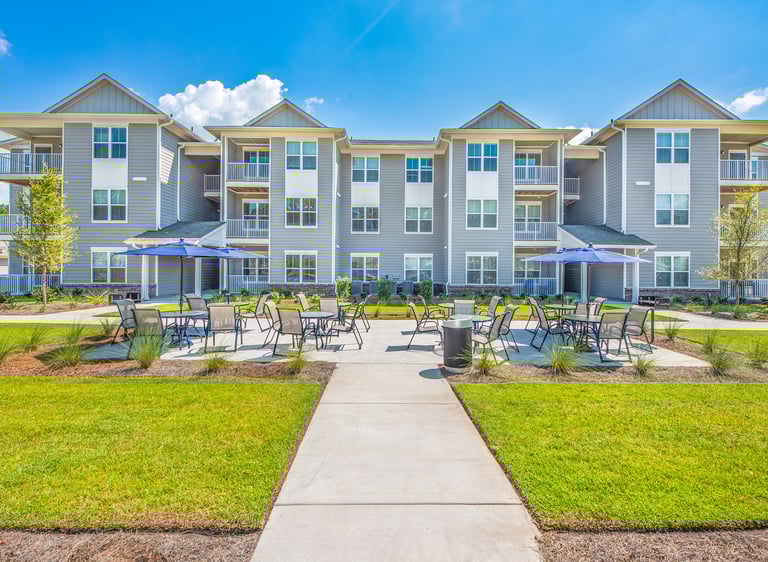 Ground-level view of outdoor patio seating with umbrellas between apartment buildings at Arrogate Village in Summerville, SC