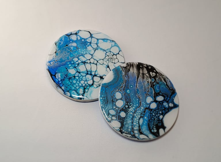 Two round acrylic pour coasters with blue, white, and black abstract cell patterns.