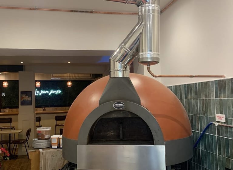 Large dome-shaped wood fired pizza oven in a modern restaurant kitchen with copper pipes.
