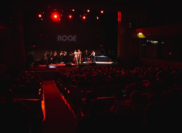 A large audience watches a live event on stage with red theater lighting