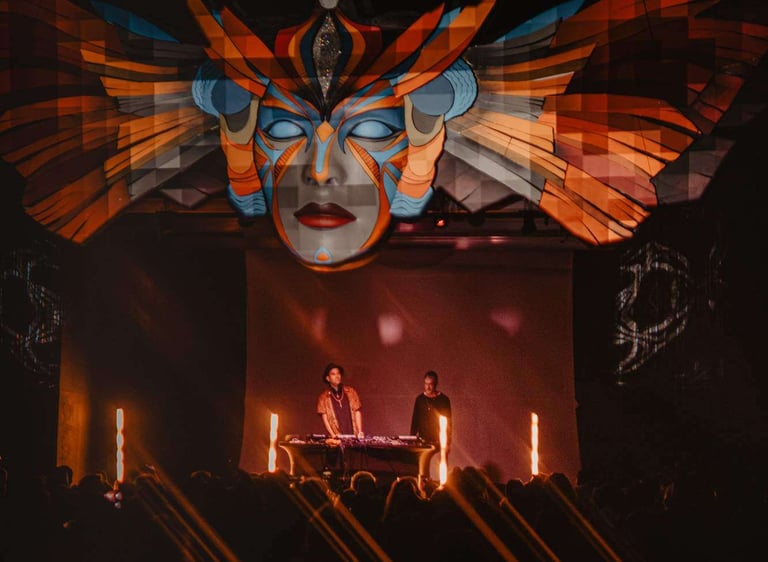 DJs performing on stage at a night music festival with a large artistic tribal mask backdrop.