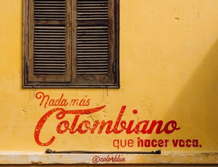 Rustic wooden shutters on a yellow wall with Colombian Spanish text and red chairs.