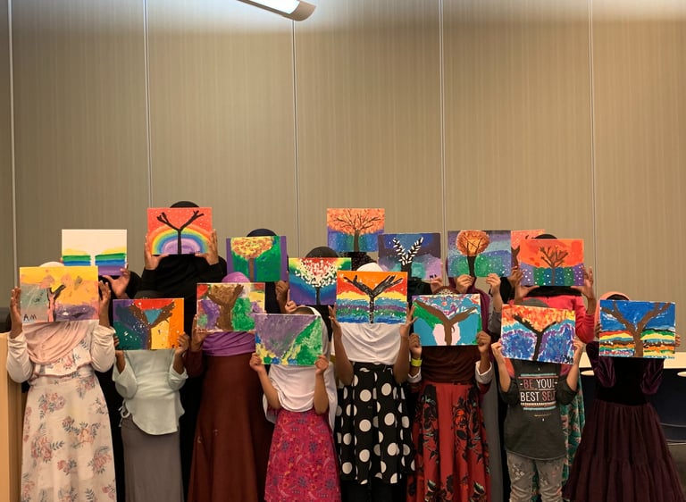 a group of Muslim girls standing in rows, holding colorful tree paintings up, hiding their faces