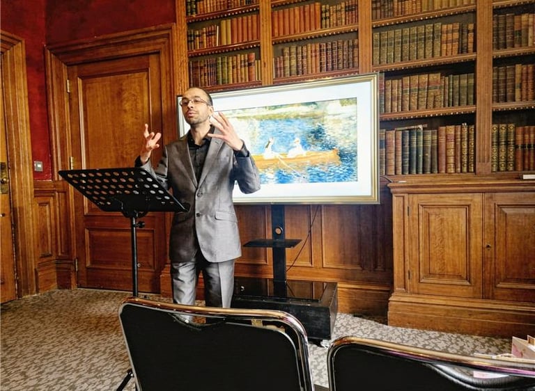 A man in a suit giving a presentation in a classic library with a digital screen showing artwork.