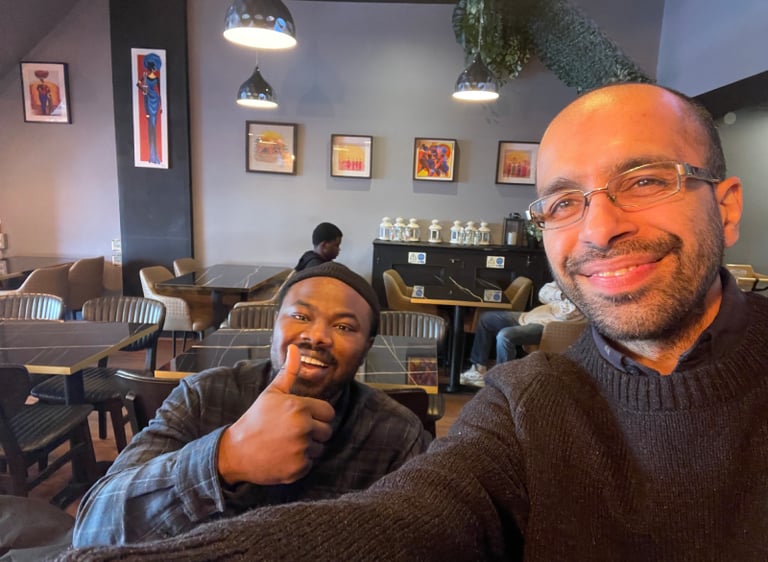 Two smiling men taking a selfie inside a modern restaurant with artwork and pendant lighting.