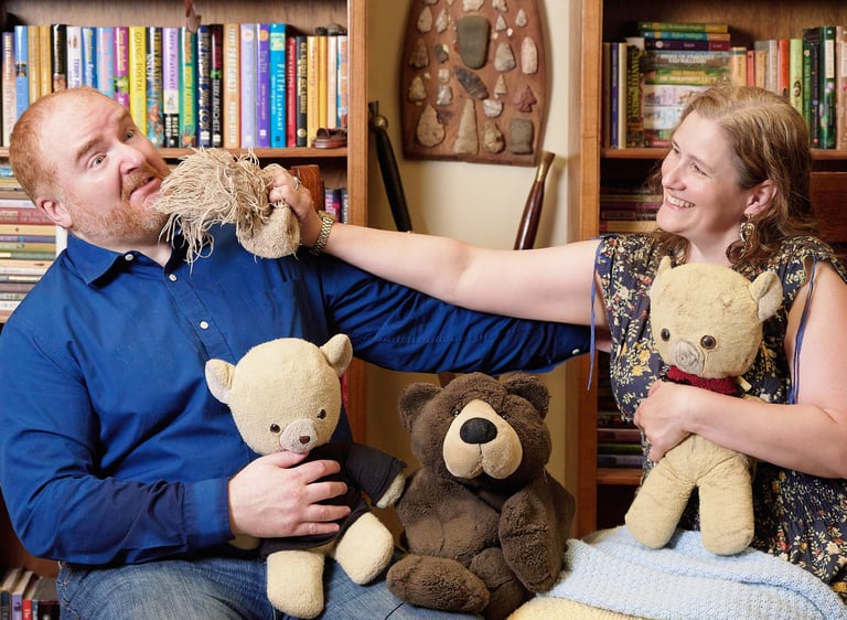 a man and woman sitting on a chair with teddy bears fighting each other