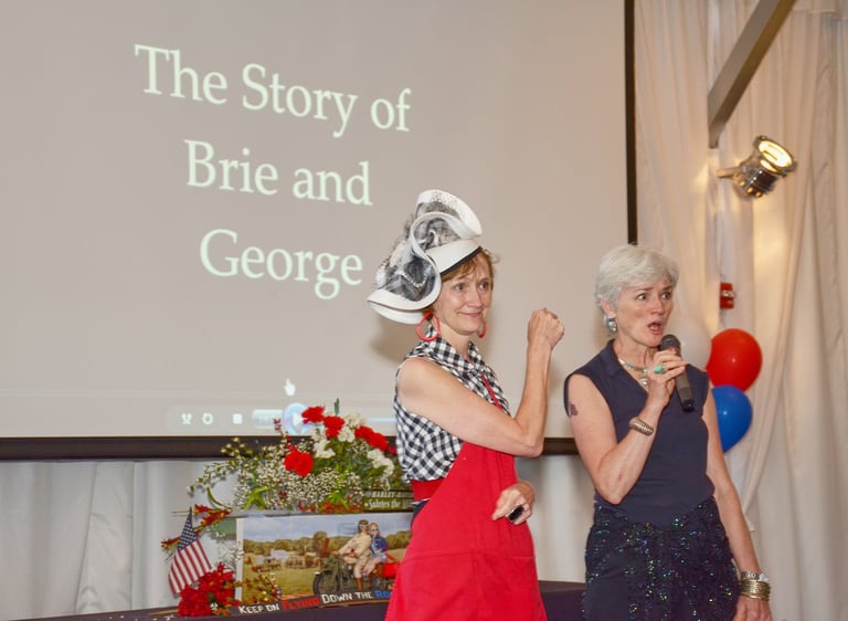 two women in a big hat and dresses standing in front of a screen speaking to the audience