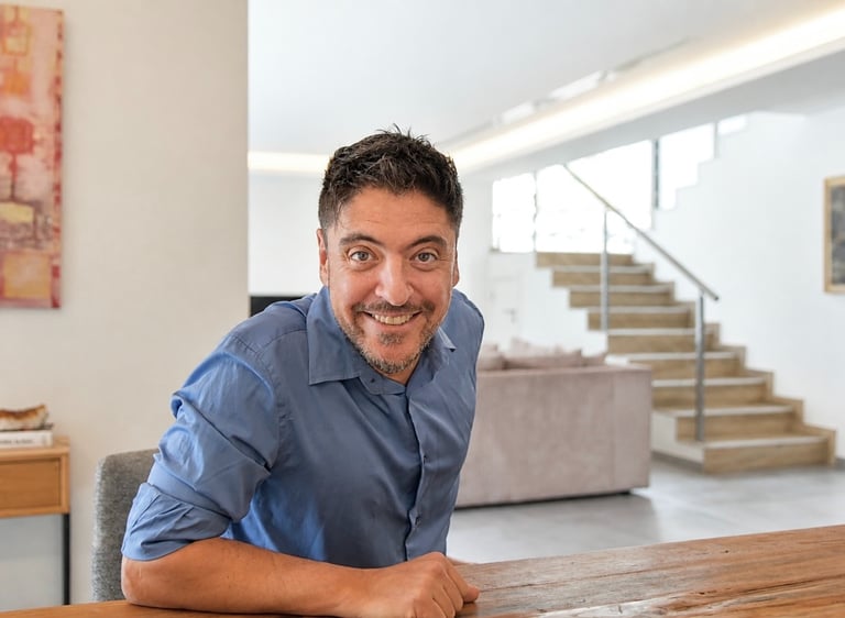 Smiling man in a blue shirt sitting at a wooden table in a bright modern home office.