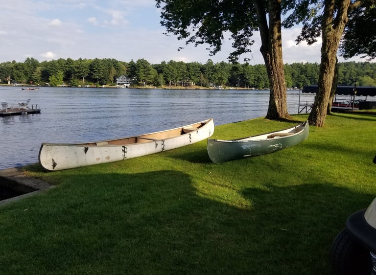 Two canoes resting on a green lawn by a peaceful lake with trees and houses in the distance.