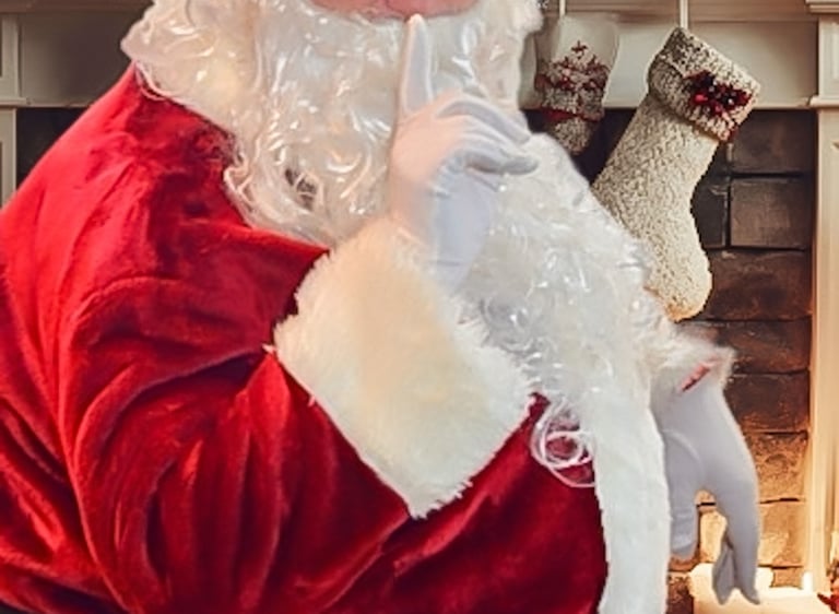 Santa Claus in a red suit making a shushing gesture by a festive Christmas fireplace.