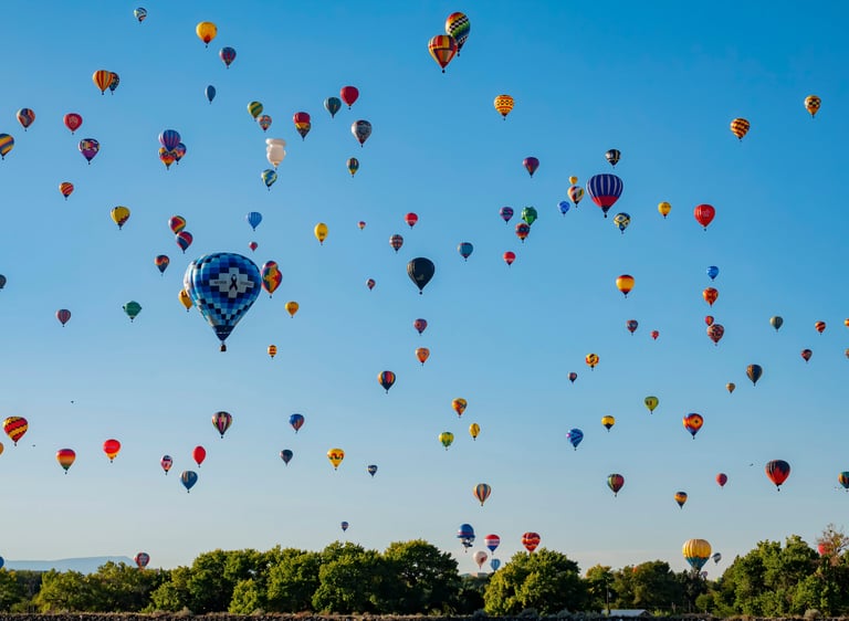 Dozens of colorful hot air balloons floating in a clear blue sky above a line of green trees.