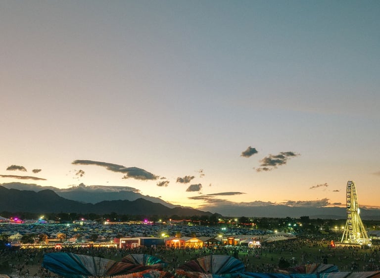 Crowds at an outdoor music festival at sunset with a Ferris wheel and colorful shade structures.