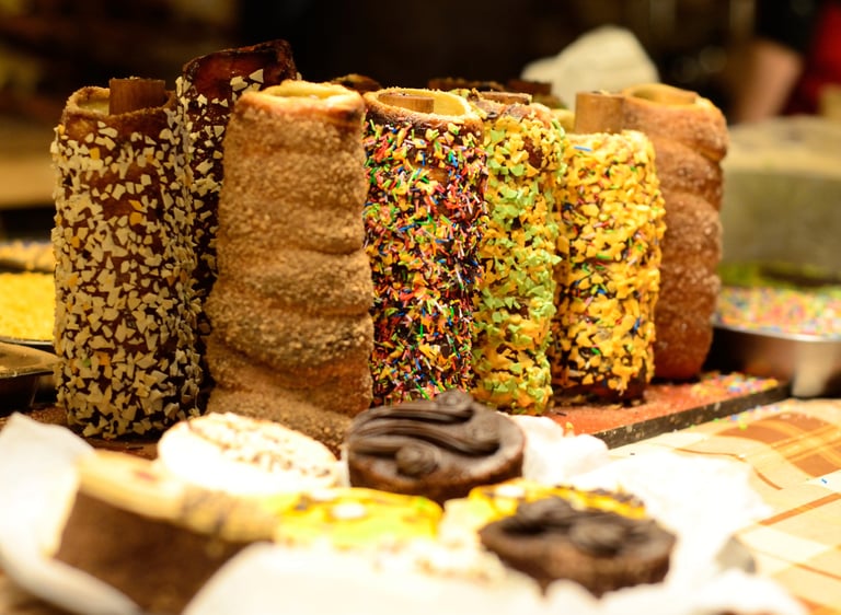 Freshly baked chimney cakes with assorted toppings like sprinkles and nuts at a pastry market stall.