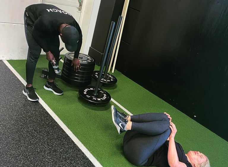Personal trainer assisting a woman performing knee-to-chest stretches on gym turf during a workout.