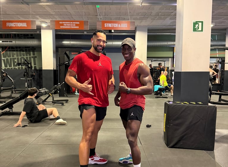 Two muscular men smiling and posing for a fitness photo inside a modern gym with exercise equipment.