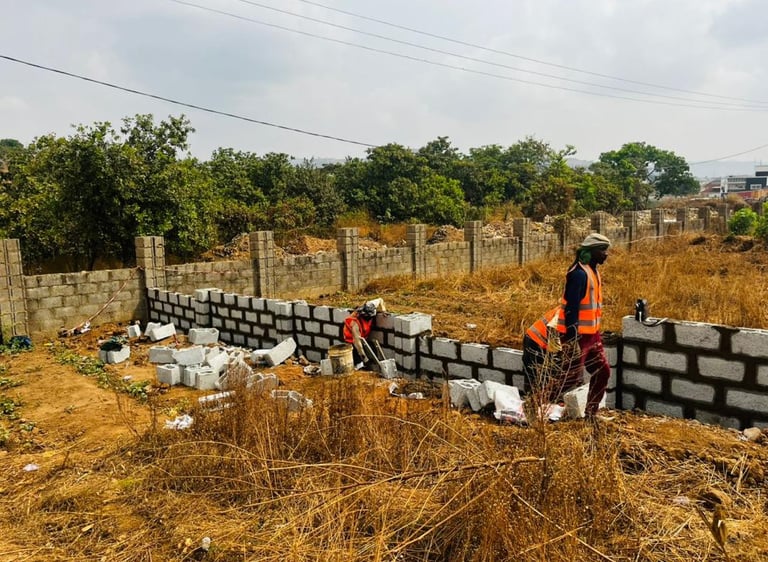 Construction workers in safety vests building a concrete block perimeter wall on a rural property.