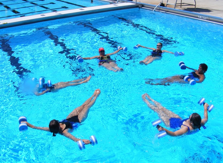 A group of women floating in an outdoor swimming pool while performing a water aerobics exercise with blue foam dumbbells.