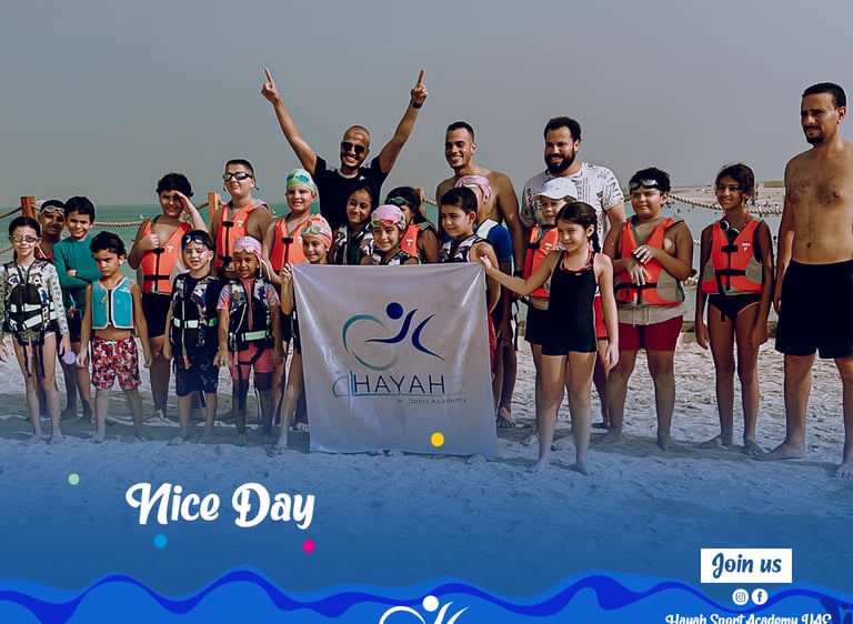 A group of children and coaches in life jackets posing with the Hayah Sport Academy banner on a beach.