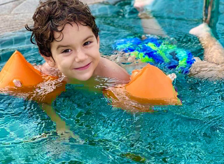 A young boy smiling in a swimming pool wearing orange arm floaties during a Hayah Sport Academy lesson.