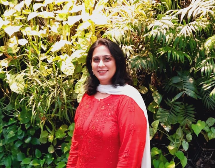 A woman in a red and white Indian ethnic suit poses against a lush vertical garden wall.