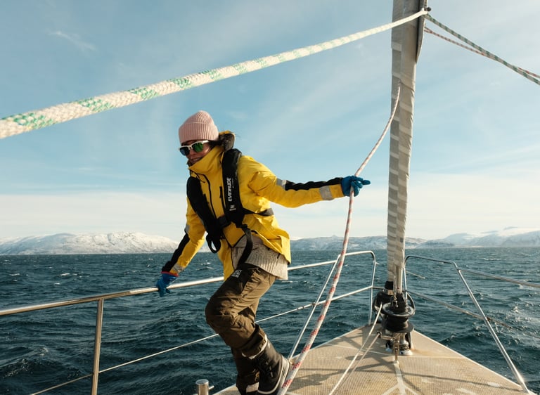 Maria på seilbåtdekk i Lofoten, gul jakke og sikkerhetsvest, vinterfjell i horisonten.