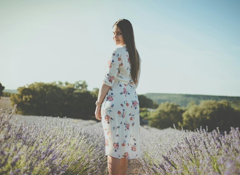 a woman in a white floral dress standing in a field of lavender. She looks at you and smiles.