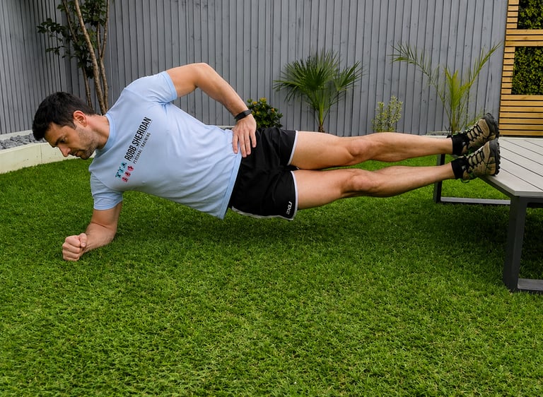 A client performing a side plank core exercise on green grass with feet elevated on a bench.
