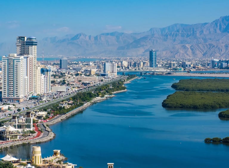 Aerial view of Ras Al Khaimah city skyline featuring skyscrapers, mangroves, and Hajar Mountains.