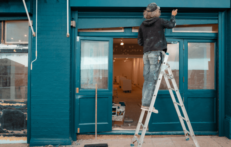 A professional painter on a ladder painting a blue storefront during a shop renovation.