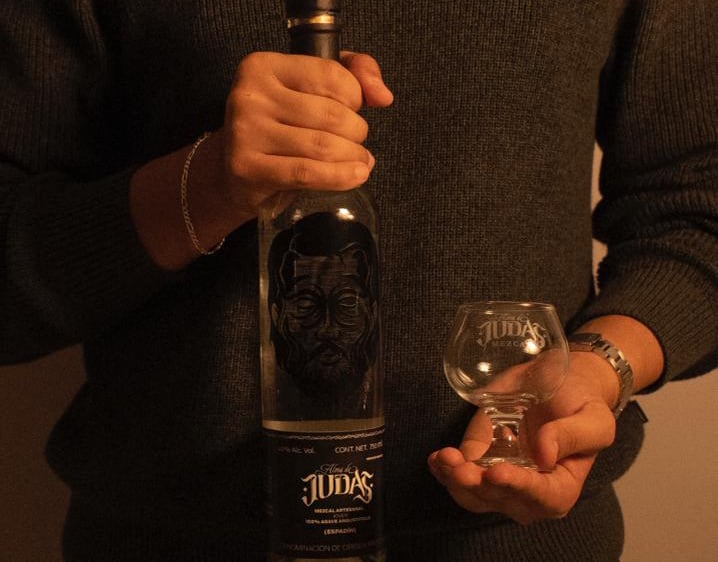 A person holding a bottle of Alma de Judas artisanal mezcal and a small tasting glass.