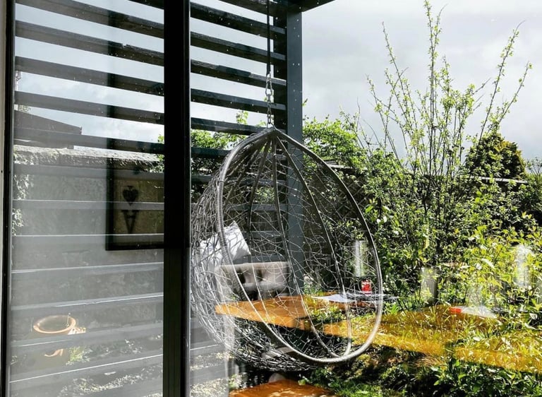 Modern hanging egg chair on a wooden deck under a black pergola in a lush garden patio.