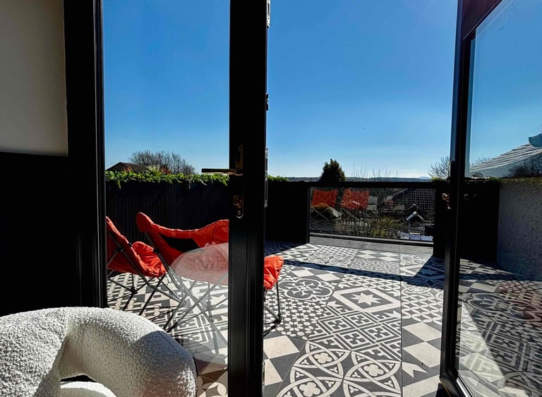 Modern rooftop terrace with patterned tile flooring, red chairs, and black French doors under a sunny blue sky.