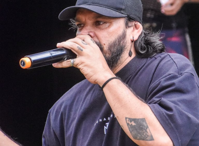 Vocalista masculino con barba, gorra negra y camiseta negra interpretando una presentación en vivo 